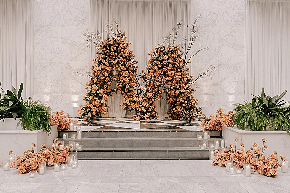 Wedding ceremony backdrop with floral ceremony arch of peach roses, greenery, and branches, candlelit on stage steps against a marble wall