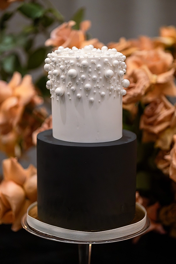 Wedding cake in black and white with matte black fondant base and cascading sugar pearls, topped with florals on a stand