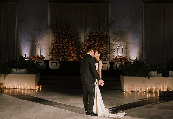 Wedding kiss portrait of bride and groom kiss in front of a candlelit head table with tall tapers, orange florals, and marble wall backdrop