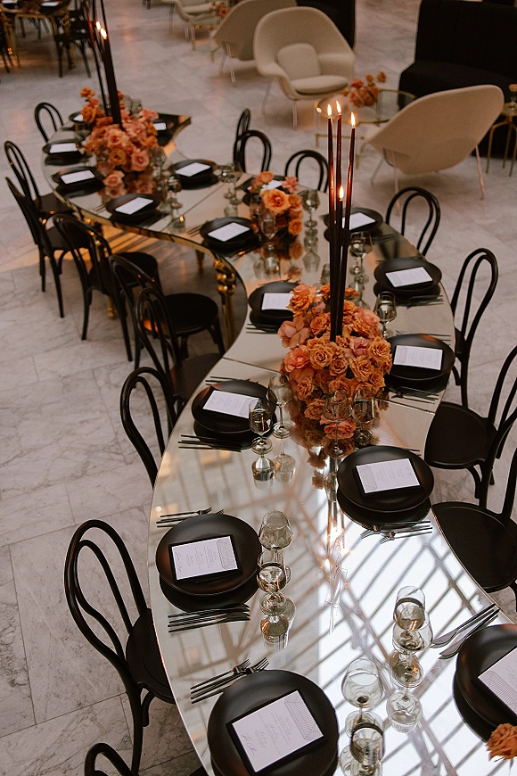 Reception tablescape with mirrored wedding table, orange floral centerpieces, tall black taper candles, black plates and menus on a marble floor venue