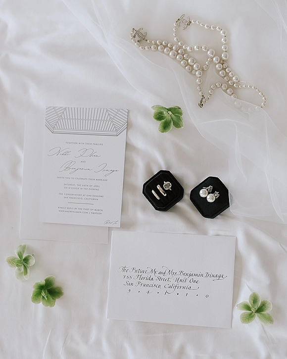 Invitation suite wedding invitation flat lay with RSVP and calligraphy envelopes, rings, veil and pearls on white fabric with green flowers