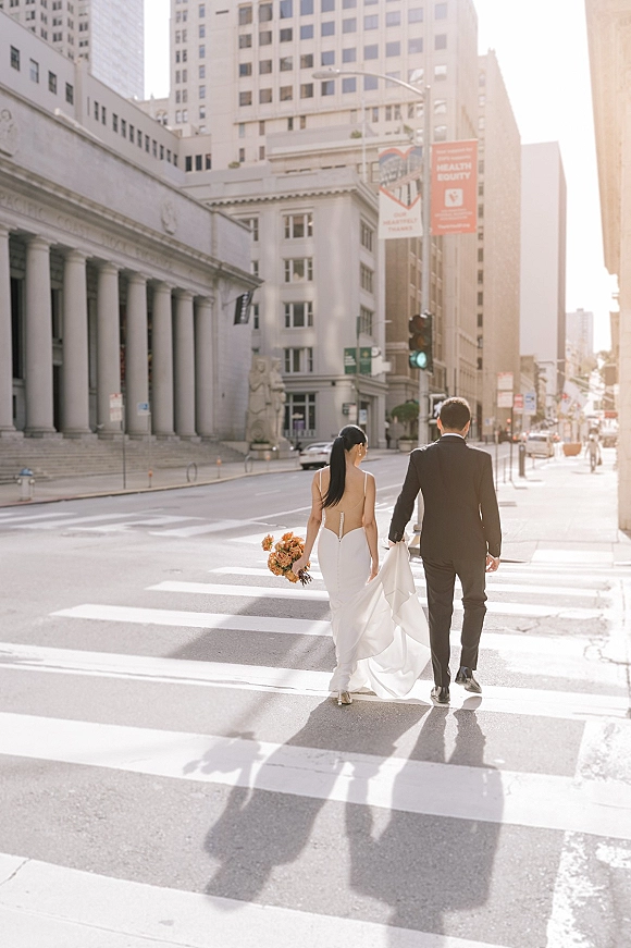 Couple portrait of bride and groom walking across a downtown crosswalk, bouquet in hand, groom lifting her dress train in sun flare