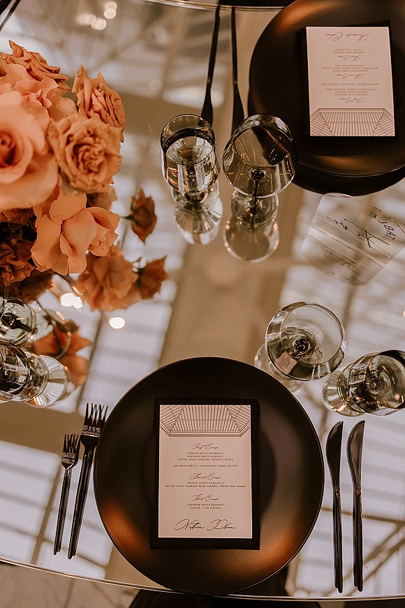 Reception tablescape with black and peach tablescape styling, black charger plates, printed menus, glassware, and a peach rose centerpiece on mirrored tabletop