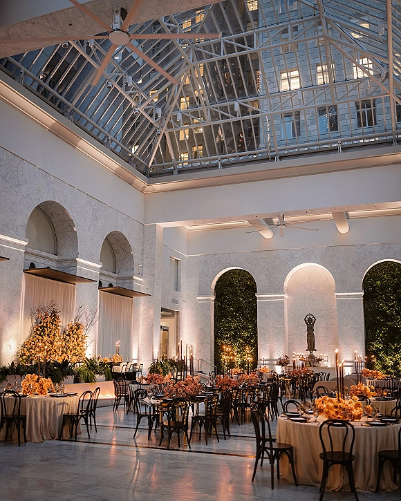 Reception tablescape with round wedding tables in cream linen, orange floral centerpieces and tall taper candles beneath a glass-ceiling atrium