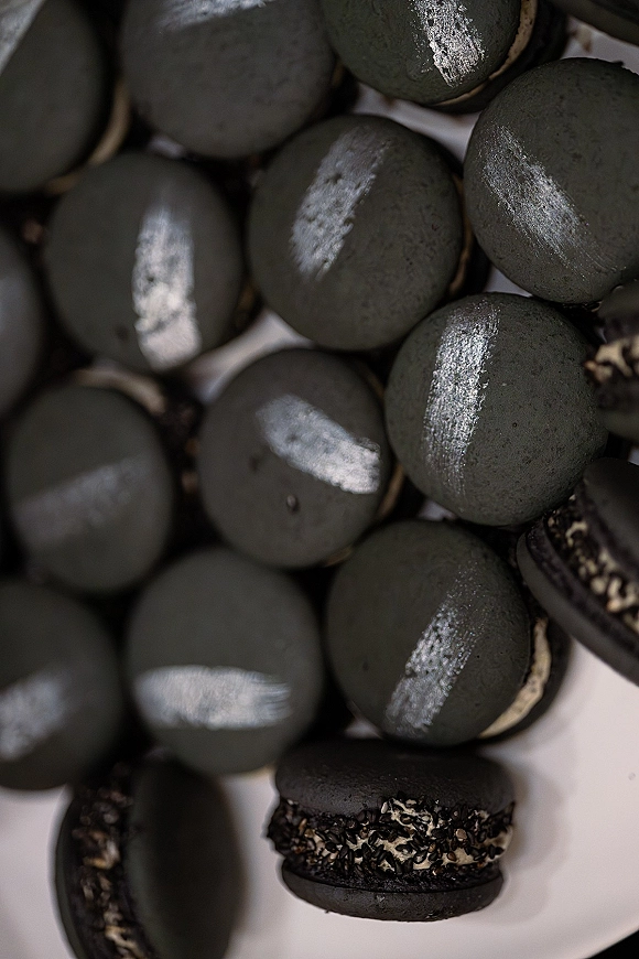 Black macarons with silver luster dust and sesame coating, showing cream filling on a white serving surface for a modern wedding dessert table
