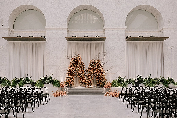 Ceremony setup for an indoor wedding ceremony with peach flower and greenery installation, white drapery, candles, and black chairs by arched windows