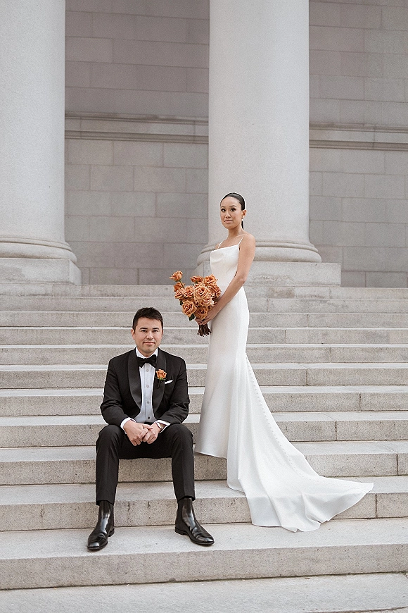 Couple portrait of bride in simple satin slip dress holding orange rose bouquet beside groom in black tuxedo on stone steps by columns