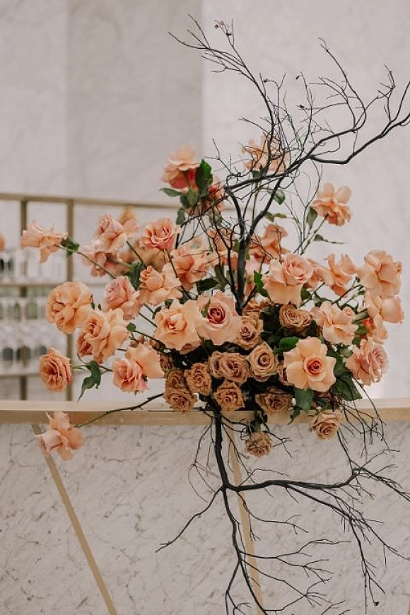 Wedding floral arrangement with peach rose centerpiece featuring blush roses and tall branches on a marble pedestal by window panes