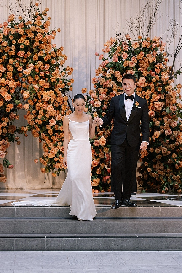 Couple portrait of bride in a sleek wedding dress and groom in tuxedo holding hands on stone steps before a rose floral wall backdrop