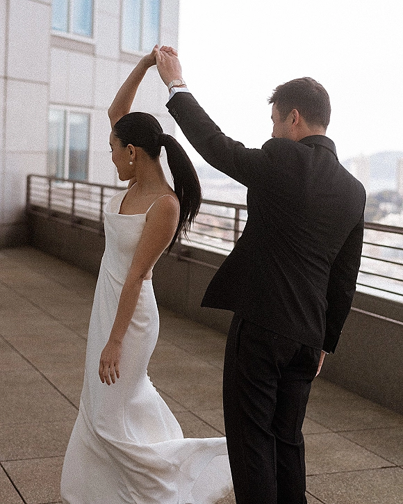 Couple portrait of the wedding couple dancing, groom in black suit leading bride twirling her satin dress on a rooftop terrace skyline backdrop