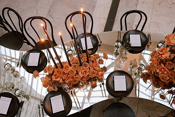 Reception tablescape with a mirrored wedding table, black plates, menus, taper candles, and peach rose centerpiece reflecting window light on marble floor