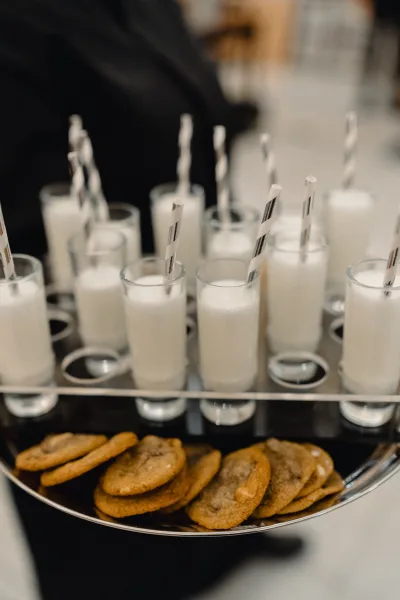Wedding dessert bar with milk shot glasses and a cookie platter on a metal tray, set in an indoor reception space with blurred guests
