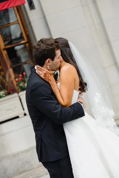 Wedding kiss portrait of bride and groom kissing on stone steps, her lace veil and henna hands on his face beside a windowed facade
