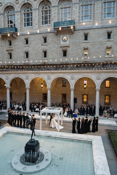 Wedding ceremony in an outdoor courtyard ceremony with bride and groom under a white chuppah, floral garland and string lights by a fountain