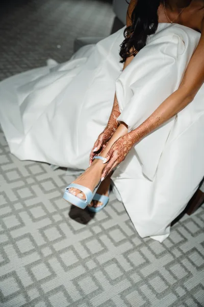 Bridal shoes in light blue wedding heels as hennaed hands slip them on beneath a white dress, on gray patterned carpet by a chair