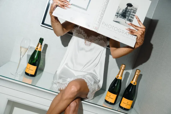 Bridal getting ready in lace bridal lingerie, henna hands showing an engagement ring beside champagne bottles and flute on a coffee table