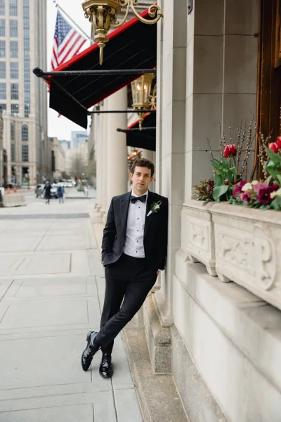 Groom portrait in a black tie tuxedo with white rose boutonniere, leaning on a stone building wall along a busy city sidewalk