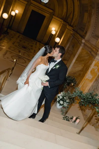 Wedding kiss portrait of bride and groom kissing on a grand marble staircase, her cathedral veil and white bouquet beside candles and greenery