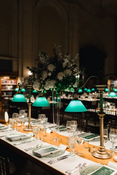 Reception tablescape at a library wedding reception with green banker lamps, tall white floral centerpiece, menus, place cards, and candles on wood table