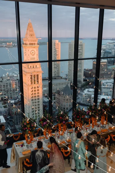 Wedding reception decor with string lights over long tables in gold linens, candles and marigold garlands by window wall at sunset skyline