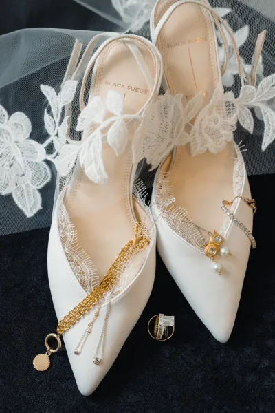 Bridal shoes, white pointed toe heels with pearl charm and rhinestone strap, styled on dark fabric with lace veil detail and rings