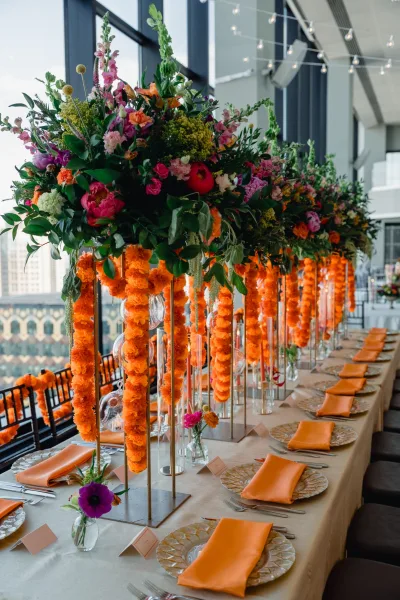 Reception tablescape with head table decor, marigold garlands, orange napkins, taper candles under string lights by loft windows and skyline