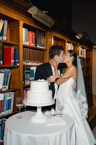 Cake cutting moment as bride and groom slice a two-tier white wedding cake, her henna hands on the knife, library bookshelves behind them
