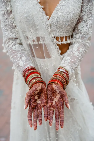 Bridal mehndi hands with intricate henna patterns and red-gold bangles, wedding ring and beaded tassels against a soft blur background
