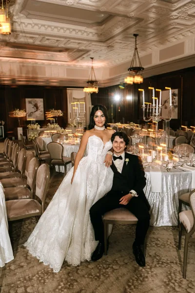 Couple portrait in a ballroom reception portrait, with a seated groom and a standing bride in a lace ballgown wearing a pearl choker.