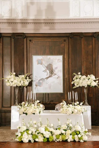 Sweetheart table decor with tall floral centerpieces, highlighted by taper candles, set against dark wood paneling and ornate plaster molding.