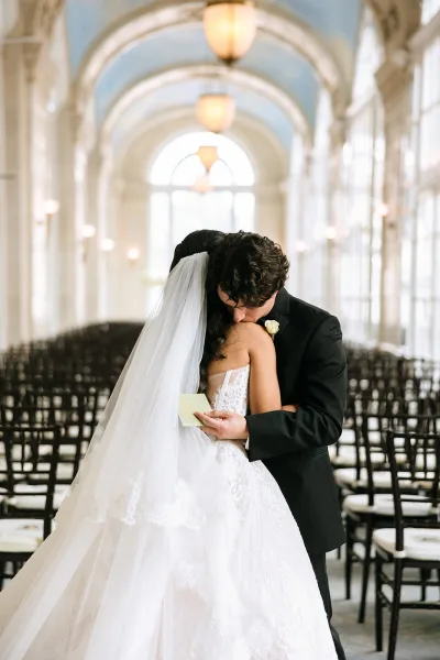 First look moment as a couple embrace, groom kisses bride with a long veil in an arched hall framed by tall windows and glowing chandeliers.