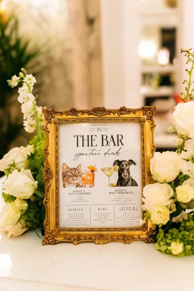 Signature drink sign and wedding bar menu featuring pet illustrations, set on a white countertop with green plants nearby.