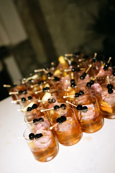 Signature cocktail display arranged for a wedding cocktail hour, poured into rocks glasses on a white countertop against a stone wall.