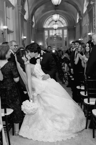 Recessional kiss during the ceremony exit in an arched hall with large windows, the bride's long veil trailing as they pause mid-aisle.