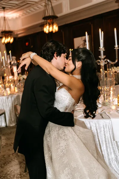 Wedding kiss portrait of the bride and groom sharing a reception kiss moment beneath ballroom chandeliers, her lace bridal gown catching the light.