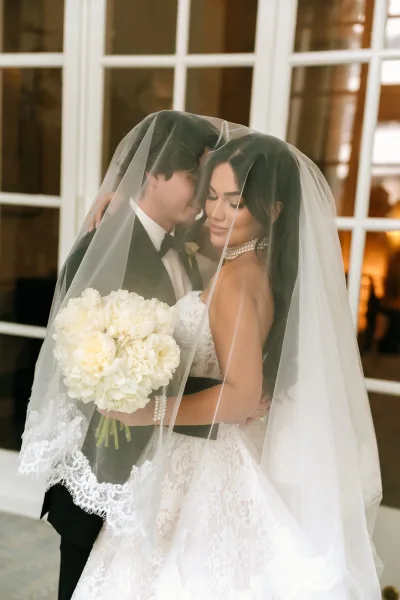 Couple portrait of a bride and groom under veil, posed closely by French doors in an airy indoor setting.