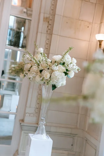 Ceremony flowers styled as a pedestal flower arrangement with white roses, set against a paneled wall with mirror and sconce.