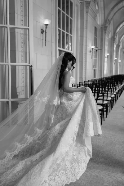 Bridal portrait of a bride in lace gown walking down an arched hallway, cathedral veil trailing amid tall windows and rows of chairs