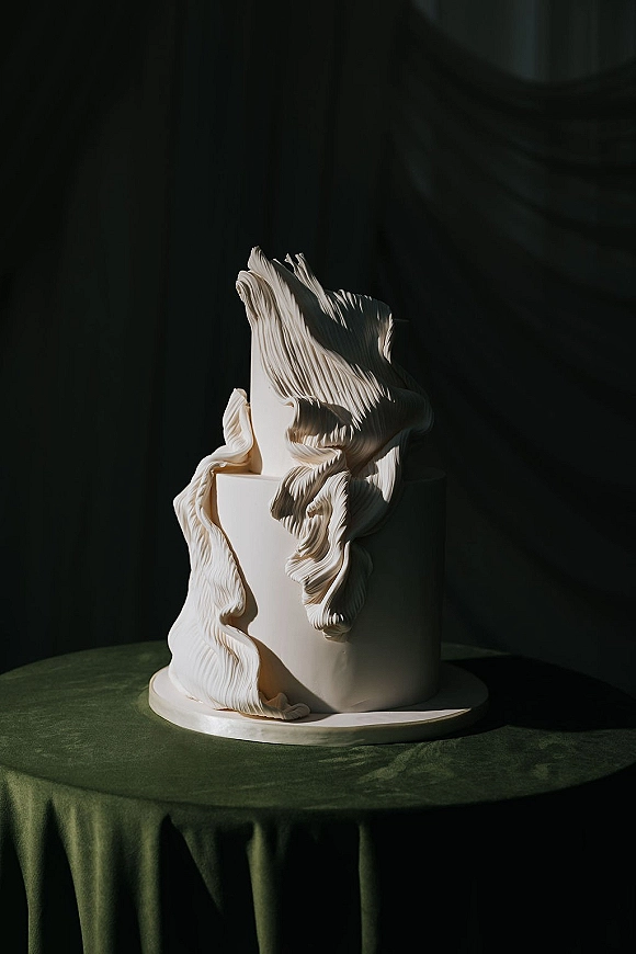 Wedding cake in white fondant with ruffled pleated texture on a cake board, set on green velvet tablecloth against a dark backdrop