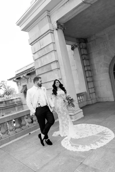 Couple portrait of a bride and groom walking hand in hand, her lace train trailing, along a terrace of stone columns at a historic building.