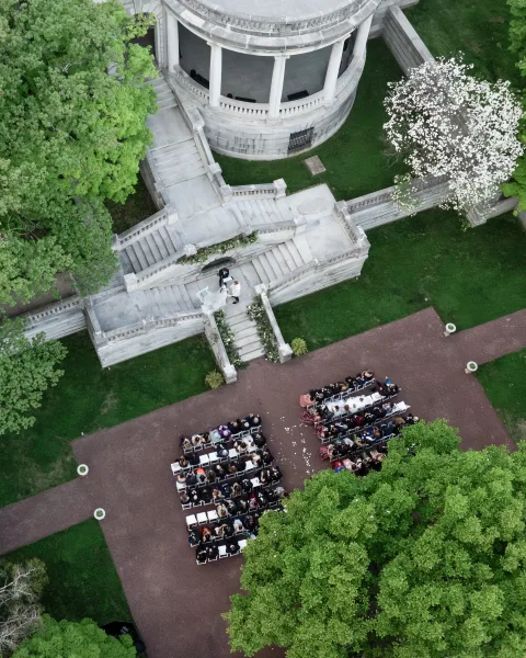 Ceremony setup with outdoor ceremony seating facing a grand staircase draped in floral garland, framed by a stone pavilion and garden trees.
