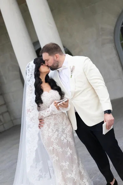 Wedding kiss portrait of a bride in lace gown and groom, framed by stone columns and an archway, her lace bridal veil softly draping.