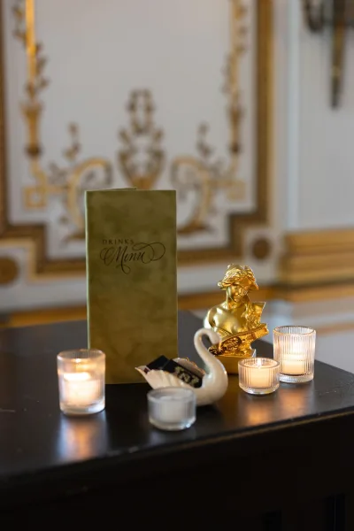 Wedding bar menu and cocktail menu sign arranged on a dark tabletop with votive candles, set against ornate wall molding with gilded accents.