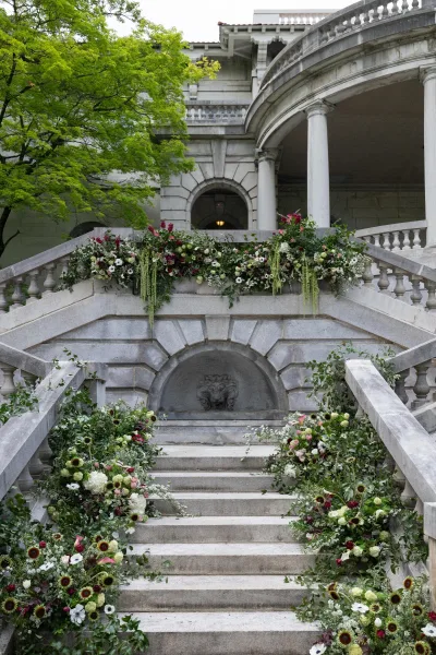 Wedding staircase flowers create a lush floral staircase installation with floral garlands on a stone stairway before a historic facade and archway.