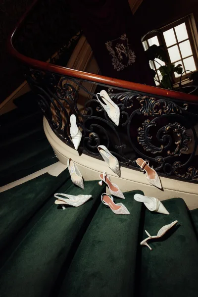 Wedding shoes, pearl-detailed bridal heels resting on a green-carpeted staircase beside a wrought iron railing and soft drapery.