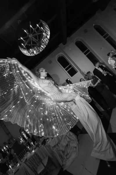 Reception entertainment featuring an LED wing dancer gliding across a checkered-floor ballroom with arched windows beneath a chandelier.