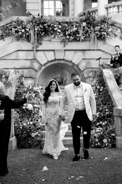 Wedding recessional of smiling newlyweds in a just married exit, as flower petals fall on a stone staircase beneath a stone arch.