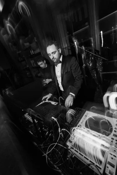Wedding DJ providing reception entertainment, mixing on turntables in a dark interior with a glass cabinet and statue.