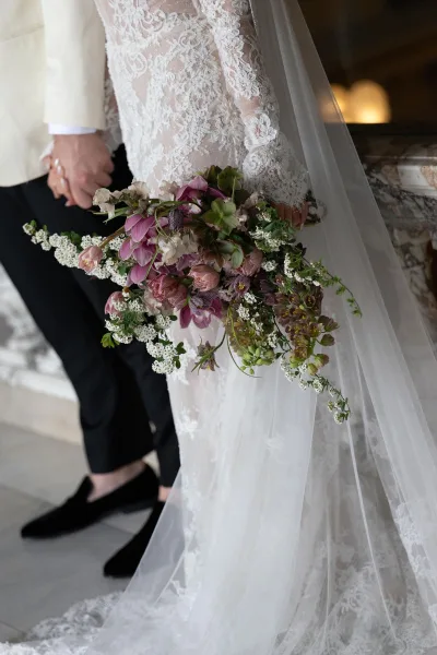 Bridal bouquet, a cascading bouquet of purple blooms and greenery, held against a lace bridal gown beside a marble balustrade indoors.