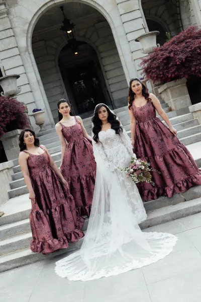 Bride and bridesmaids pose, the bride in a long sleeve wedding dress holding a blush and green bridal bouquet on stone steps by an arched doorway.